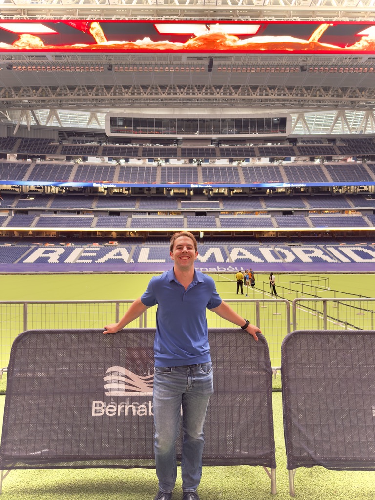 Zachary smiling in a blue polo, leaning on barriers in front of the pitch at the Santiago Bernabéu stadium